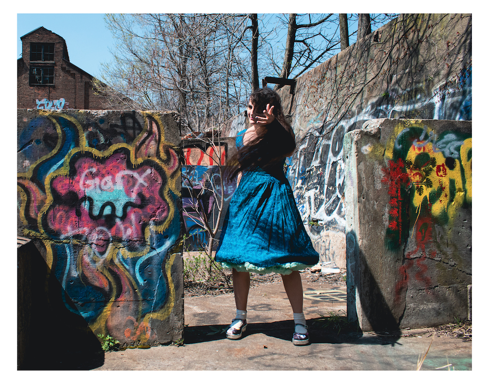 Woman reaches forward through her blowing hair in invitation, surrounded by crumbling, graffiti-painted walls