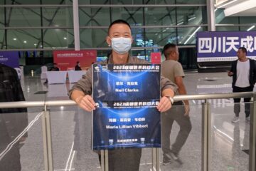 A man in an airport holding two signs - one for Neil Clarke and one for Marie Vibbert.