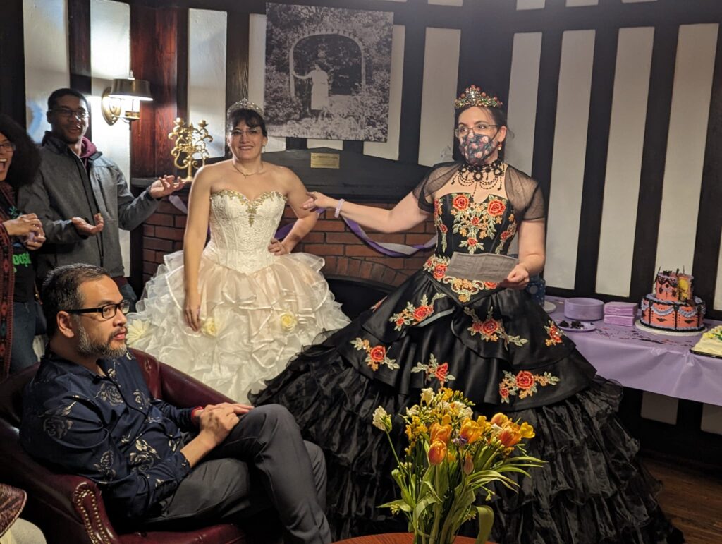 two ladies in ball gowns in front of a big cake and a crowd of friends.