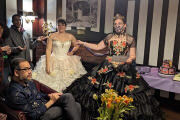 two ladies in ball gowns in front of a big cake and a crowd of friends.