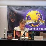 Marie at a table with her books on it with a banner behind her with the Dragon Con logo and the words "Fantasy track"