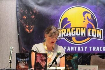 Marie at a table with her books on it with a banner behind her with the Dragon Con logo and the words "Fantasy track"