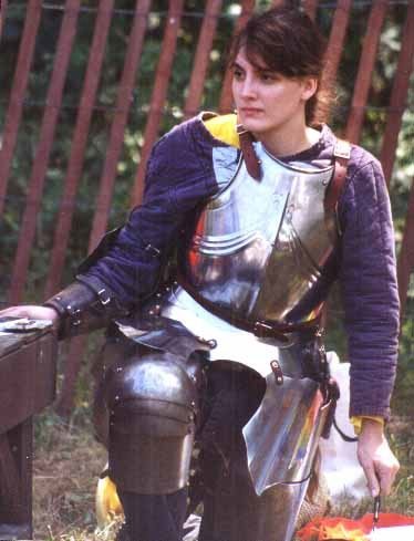 A younger Marie at a renfair, in armor with a calligraphy pen in hand. Picture somewhat marred by duct-tape holding her gambeson shut and snow fence in the background